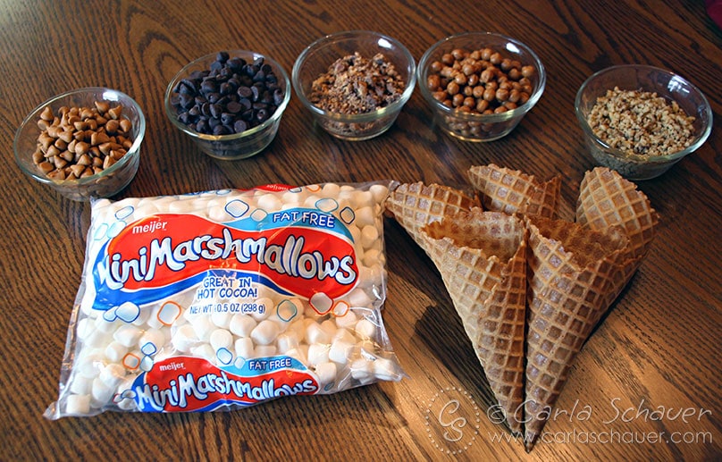 waffle cones, a bag of marshmallows and 5 small glass bowls holding candy and nuts sitting on a wooden table.