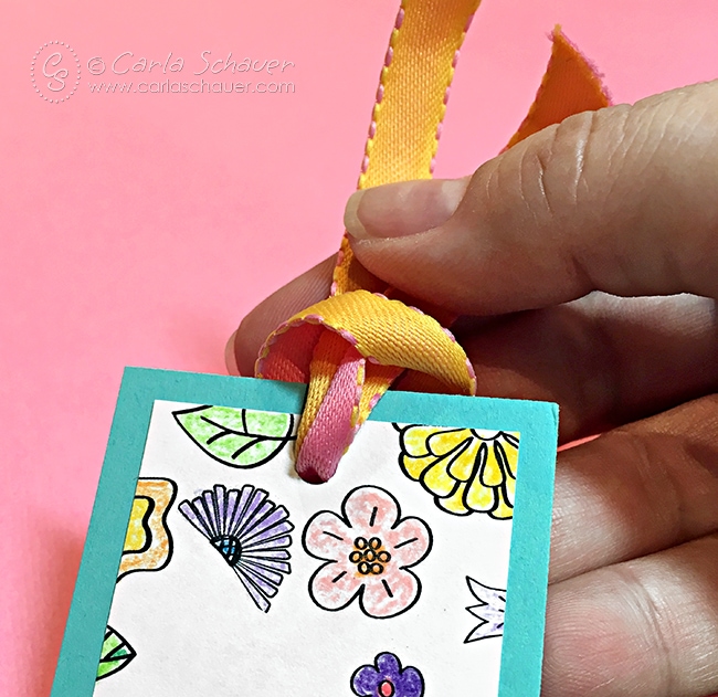 The top of a multicolored floral bookmark with hands tying orange ribbon. Pink background.