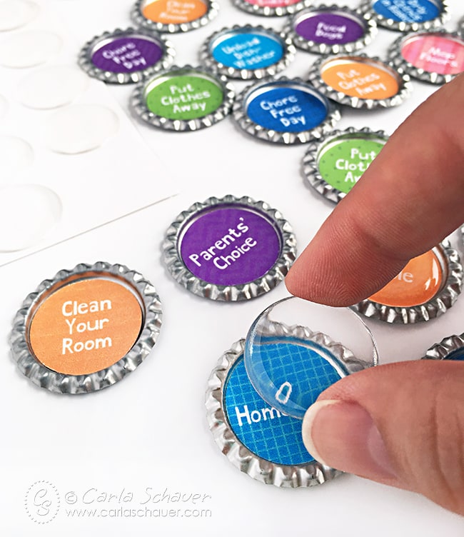 fingers placing clear epoxy circle over center of blue patterned paper inside flattened bottlecap. other colorful centered bottlecaps in background.