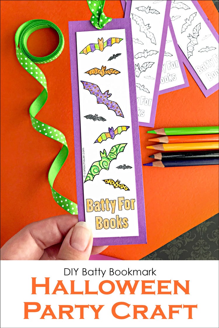 Bat bookmark colored orange, green, purple and yellow, with purple border and dotted green ribbon. Held over orange background with ribbon spool, colored pencils and more bookmarks. Text overlay reads "DIY Batty Bookmark, Halloween Party Craft"