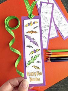 Colorful bat bookmark with colored pencils, ribbon, and uncolored bookmarks in background.