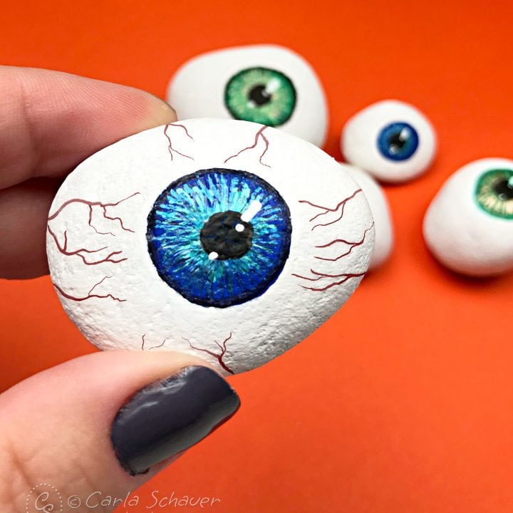 Halloween painted rock eyeball held in front of other painted eyeball rocks on orange background
