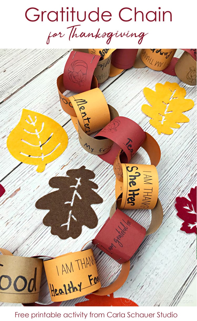 Interlocking fall colored paper strips making chain of gratitude. With felt leaves on white wood table. Red text on white at top reads "Gratitude Chain for Thanksgiving"