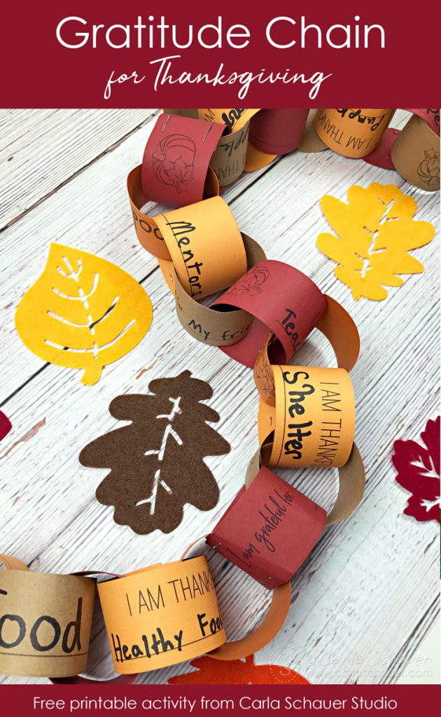 nterlocking fall colored paper strips making chain of gratitude. With felt leaves on white wood table. white text on red stripes at top and bottom read "Gratitude Chain for Thanksgiving" and "free printable actiity from Carla Schauer Studio"