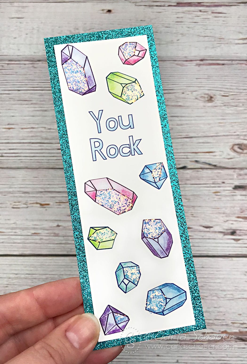 Crystal patterned glittered bookmark glued to teal glittered cardstock border, held in front of white wood background.