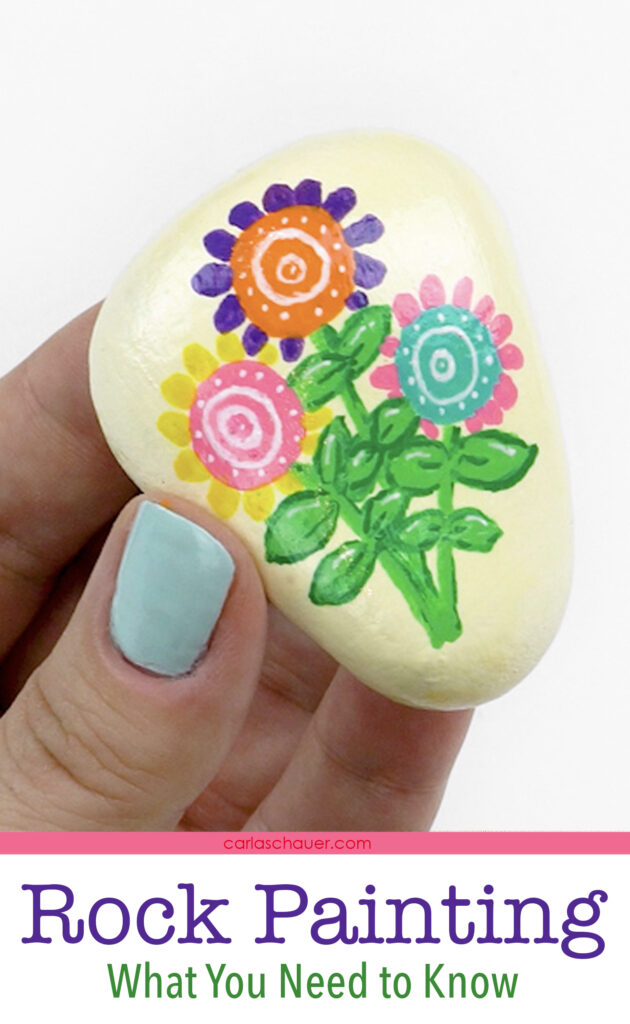 Hand holding cream painted rock with colorful flower design.