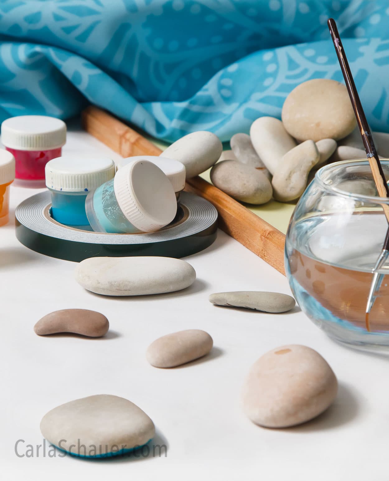 Smooth rocks, paint jars, and fishbowl with paintbrushes in water. Rock painting scene with teal fabric background.