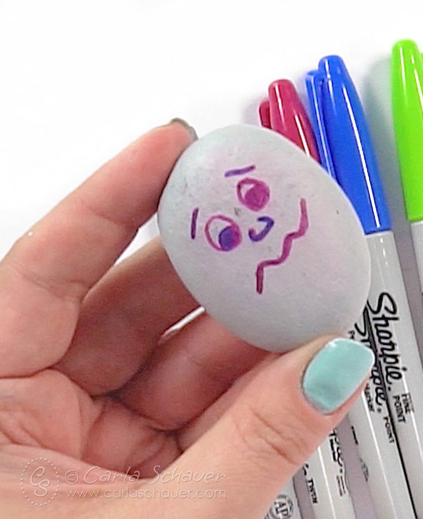 Hand holding rock with smeared sad face drawing, with markers in background.