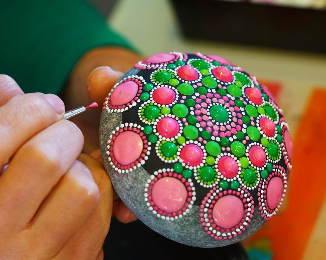 Hand holding brush is painting pink dots onto a round gray rock, working on a pink and green mandala pattern.