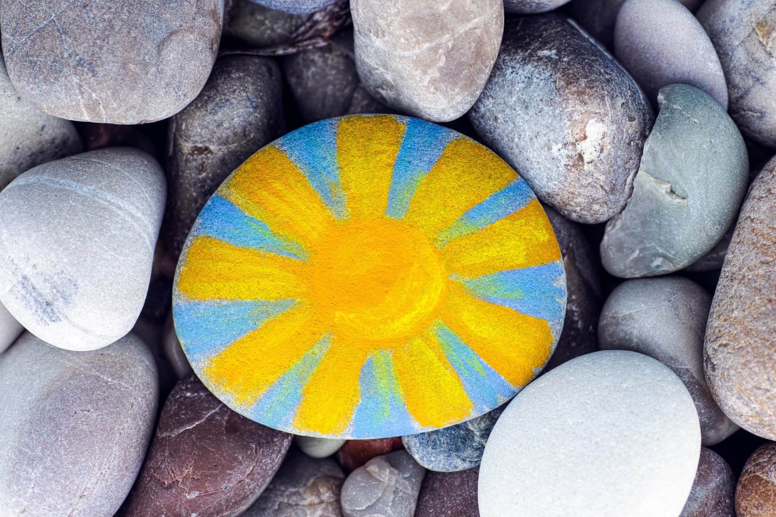 Oval rock painted blue with a yellow sunshine, sitting among unpainted rocks.