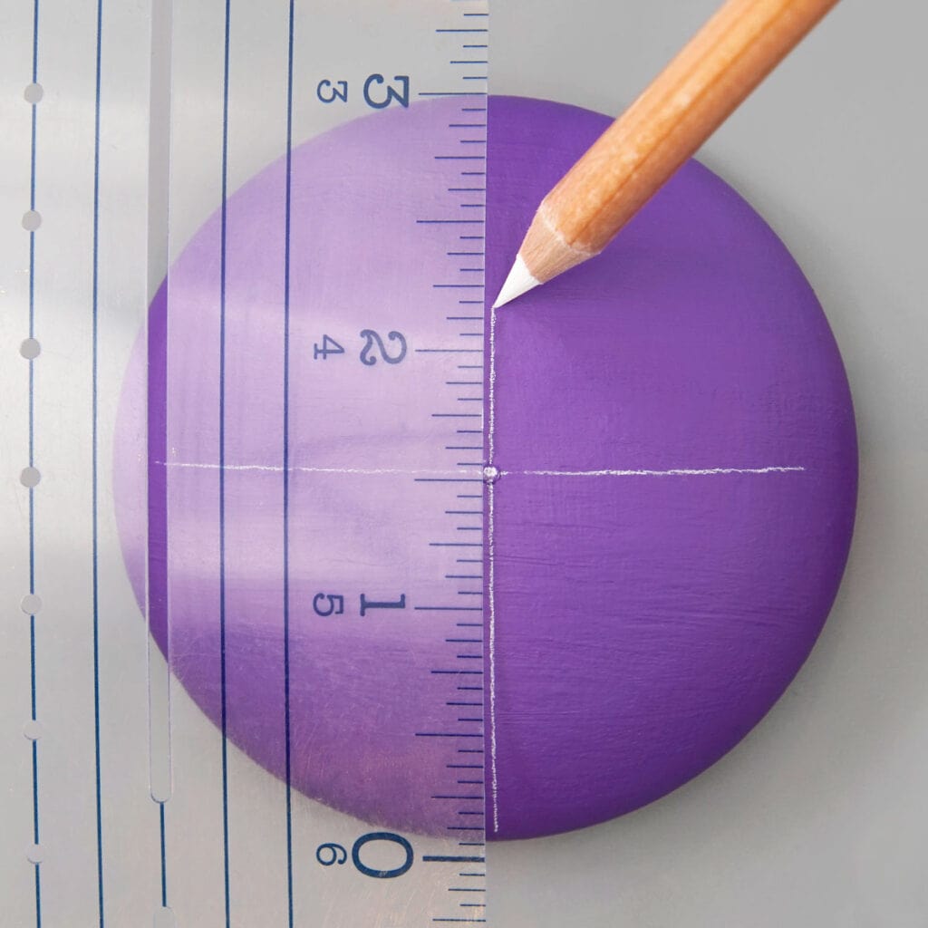 Round purple painted rock on gray background. A wood pencil with white tip uses a clear ruler to make straight lines forming a + on the rock.