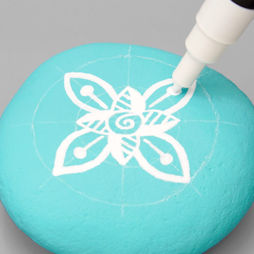 An aqua painted stone with white chalk grid markings. A white paint marker is drawing a mandala on the stone.