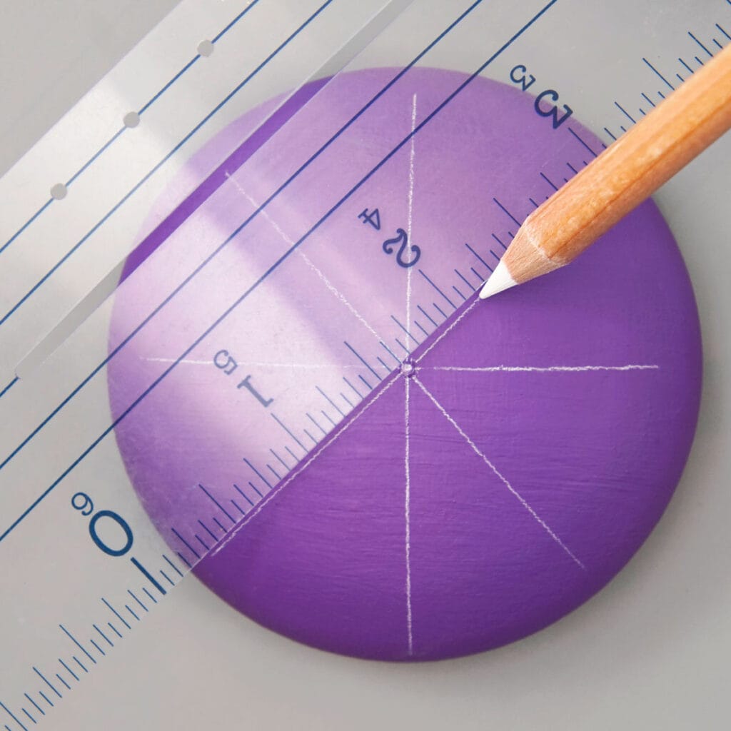 purple painted rock on a gray background. a wood pencil with white tipe uses a ruler to draw an X on the rock.