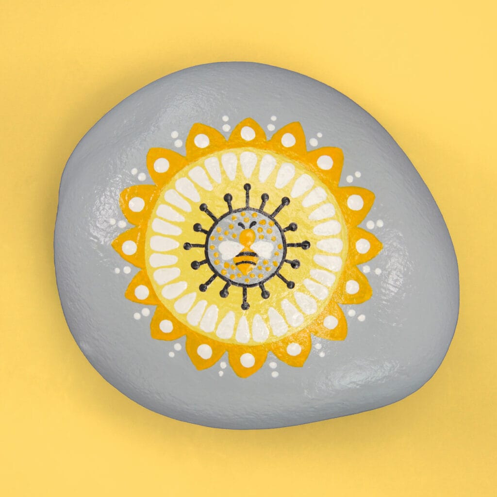 Gray painted stone on a yellow background. Stone is painted with a yellow, white, and black mandala with a small bee in the center.