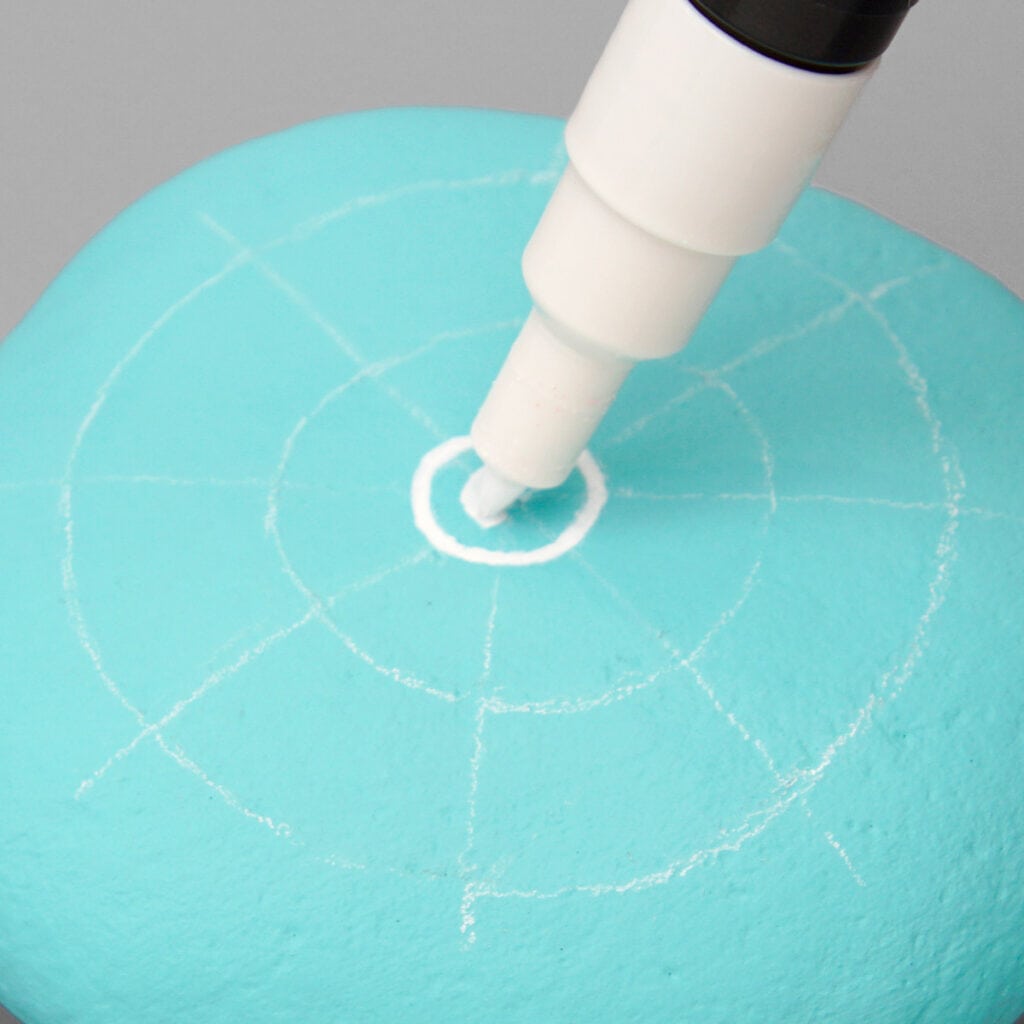 An aqua painted rock with a mandala grid marked in chalk pencil. A white paint marker is drawing a center dot surrounded by a circle.