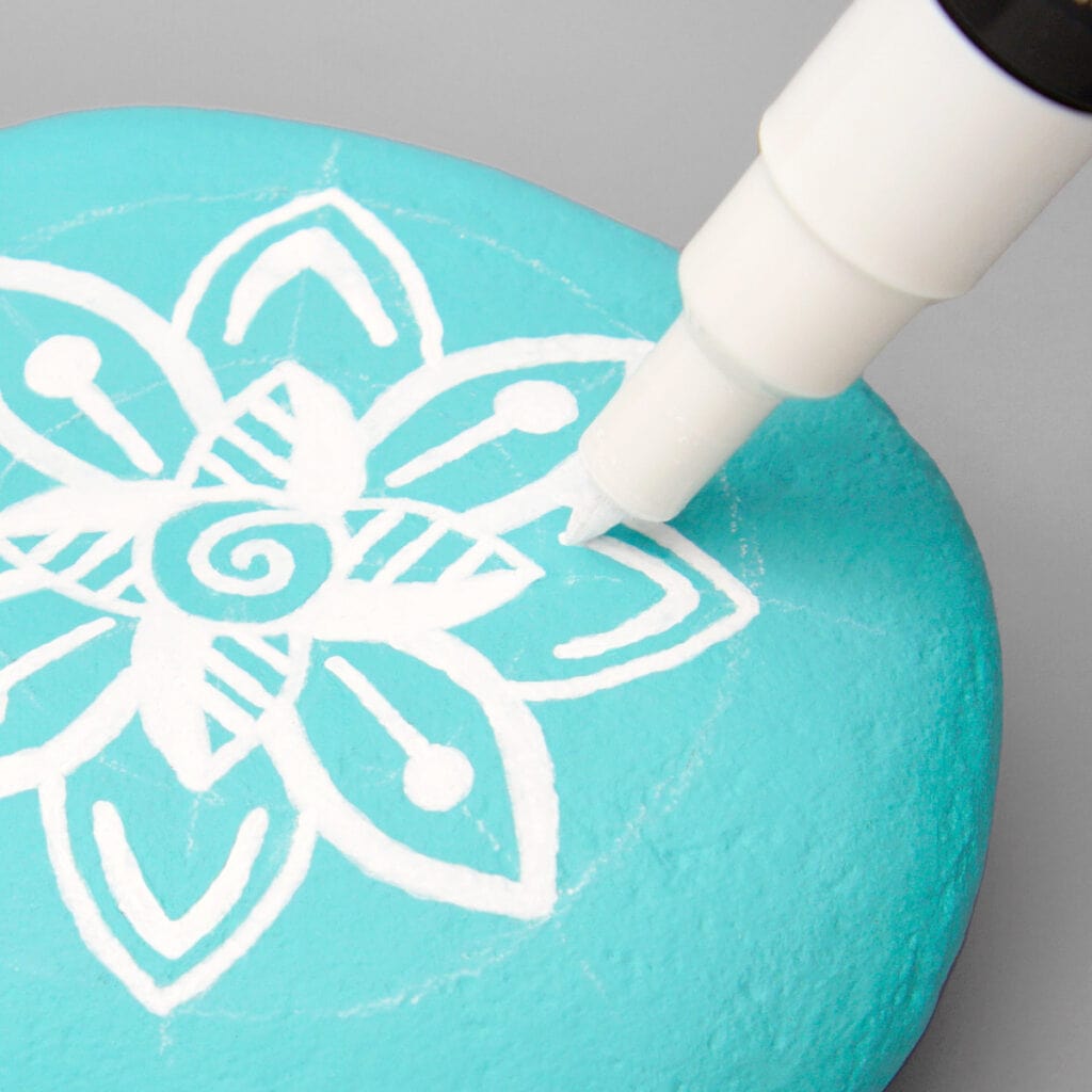 An aqua painted rock with a white chalk mandala grid. A white paint marker is drawing V-shaped accent marks inside a mandala petal.