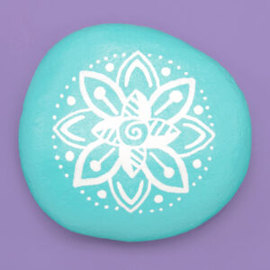 A round teal painted rock on a purple background. The teal rock is painted with a white mandala design.