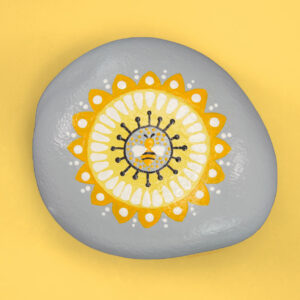 A gray rock on a yellow background. The rock is painted with a yellow, white, and black mandala design with a bee in the center.