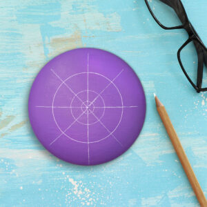 Purple painted circle rock with a white mandala grid drawn on it. Rock is lying on a distressed blue background next to a white chalk pencil and black eyeglasses.