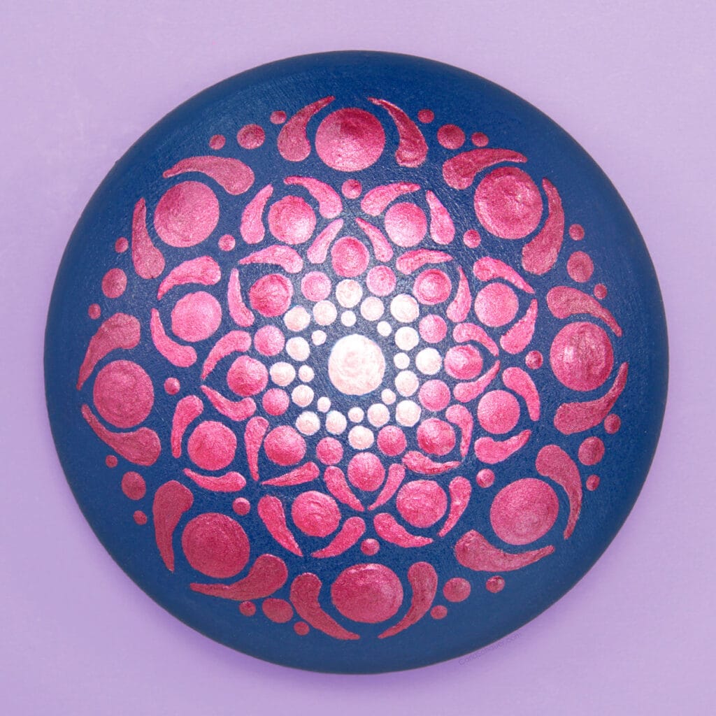 Navy blue rock on a purple background. Rock is painted with light pink to dark pink shimmery painted dot mandala.