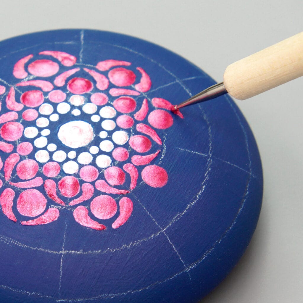 A wood dotting tool with a small silver tip drags fuchsia paint. The paint makes a comma shape in the 3rd circle of a pink ombre mandala on a navy blue rock.