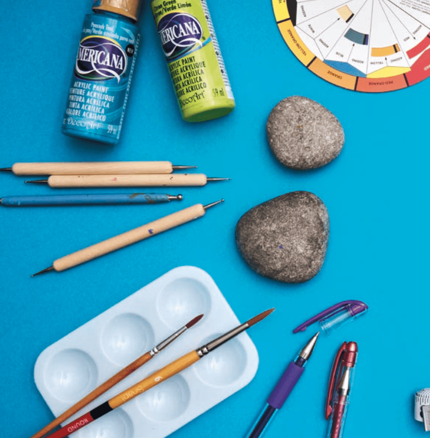 A blue background filled with rock painting supplies, including gray rocks, a color wheel, blue and green paint, paint brushes, and a white paint palette.