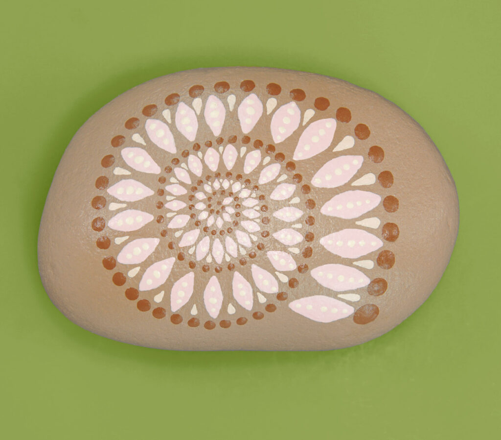 An oval tan rock on a bright green background. A spiral mandala is painted in shades of pink, cream, and brown.