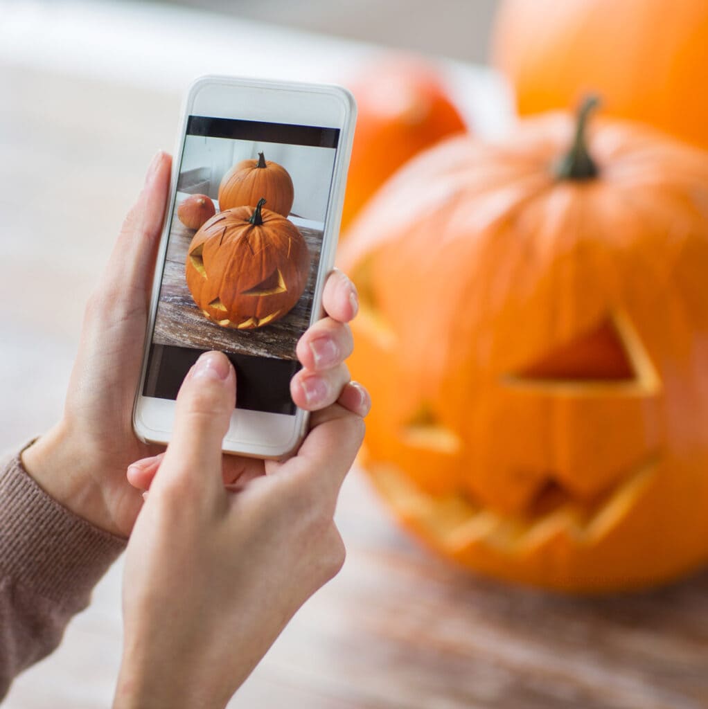 Hands holding a mobile phone, taking a photo of a carved Halloween jack-o-lantern.