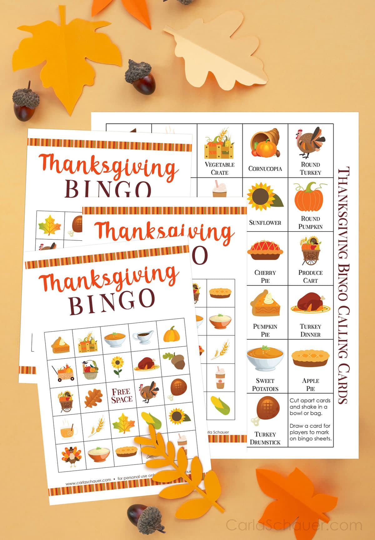 3 Thanksgiving bingo cards and 1 calling card printed pages with colorful fall icons. Printed pages are lying on a pale orange background with fall leaf paper cutouts and acorns.