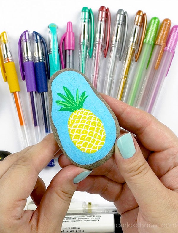 hands holding a pineapple painted rock in front of a fan of gel pens