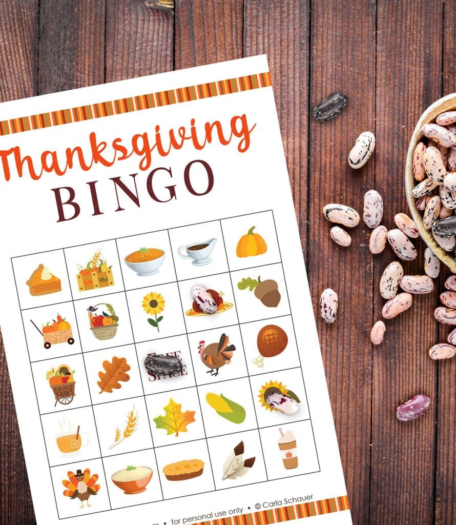 Printed Thanksgiving Bingo Game Board on a brown wood table background, next to a a bowl of dried beans. Several beans are scattered on the table, and marking the game board. Each bingo card is filled with colorful fall icons and text reading "Thanksgiving BINGO" across the top.