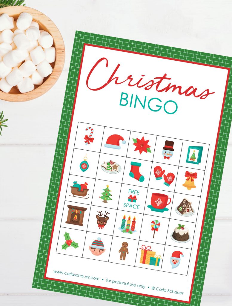 Printed Christmas bingo card lying at an angle on a white wood background next to a bowl of marshmallows. The card has colorful christmas pictures and a green border, with text at the top that reads "Christmas Bingo"