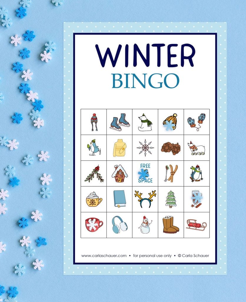 A printed winter bingo card on a light blue background with small light blue and white plastic snowflakes. The bingo card has a light blue border and a 5x5 grid of cute winter icons. Blue text at the top of board reads "Winter BINGO"