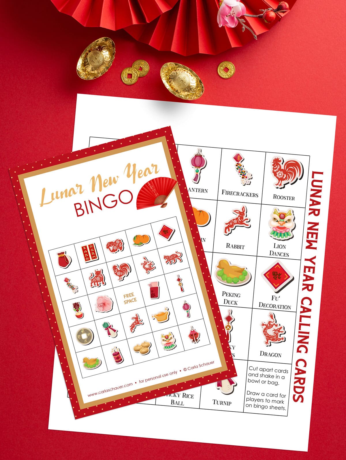 Printed Lunar New Year Bingo Card, white with red border and grid with 25 holiday icons. Card is lying on printed calling card page with same icons. Background is red with gold trinkets and red fans.
