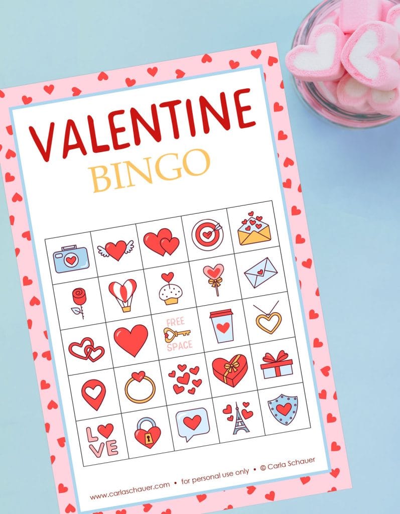 Printed Valentine Bingo card, white with a pink/red heart border and a 5x5 grid of valentine icons. Red and yellow text at top reads: "Valentine Bingo." The card is lying on a blue background next to a bowl of pink heart marshmallows.